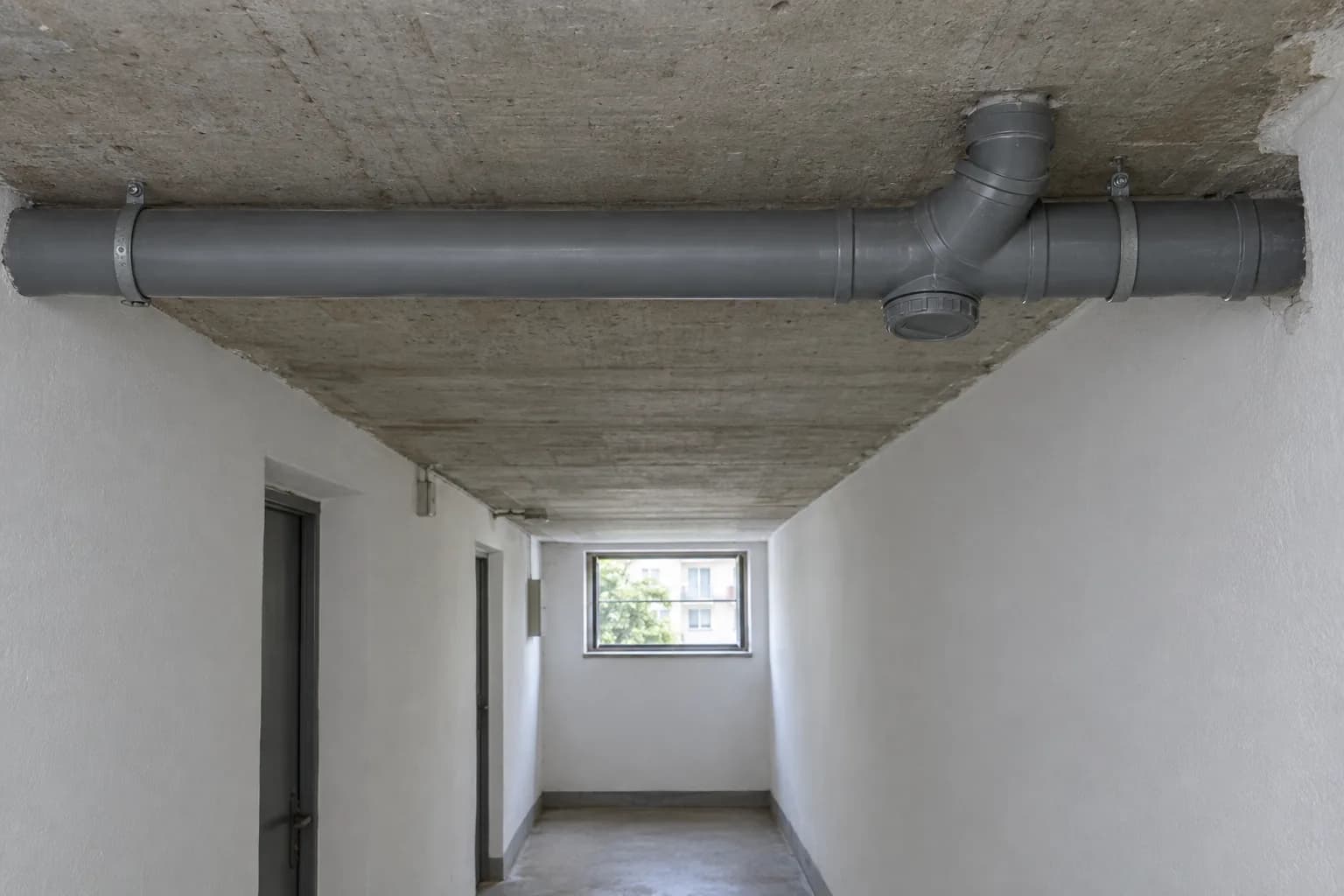 Tubo de esgoto cinzento horizontal no teto de betão de um corredor de cave, com uma derivação e uma tampa de limpeza