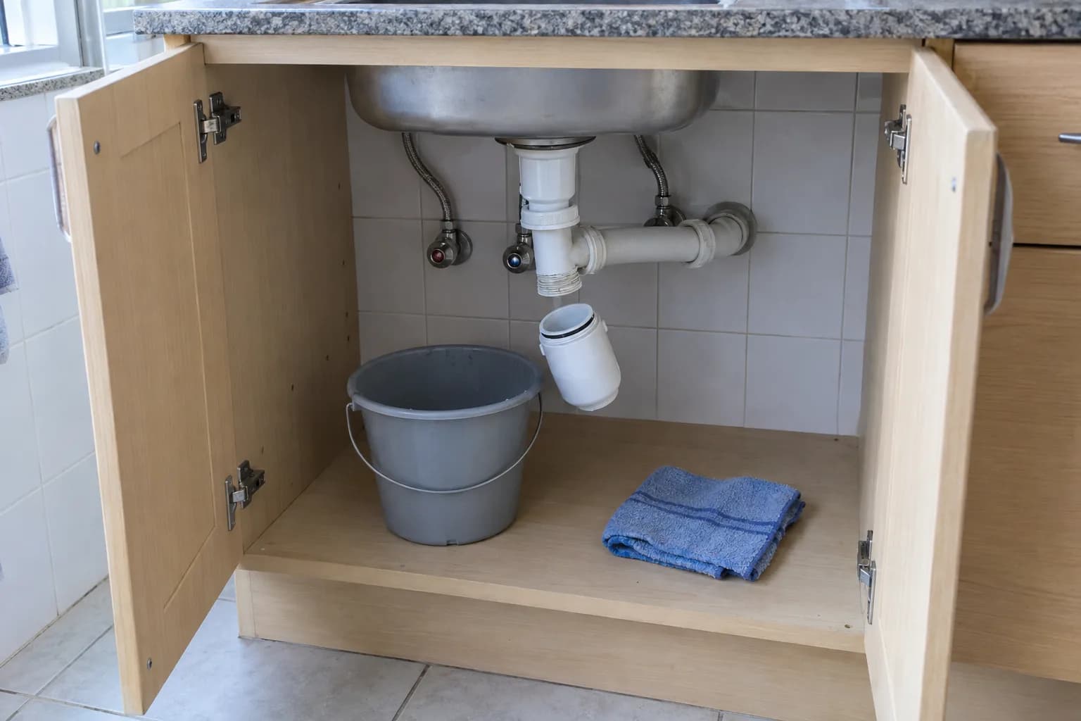 Armário debaixo do lava-loiça com as portas abertas, o sifão de plástico branco desenroscado, um balde e um pano dobrado