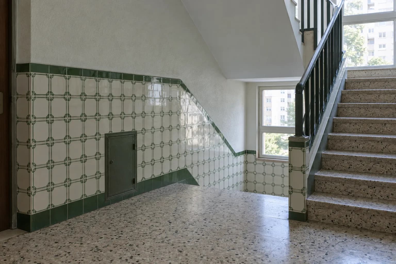 Patamar de uma escada de prédio com painel de azulejos verdes e brancos e uma pequena porta de serviço fechada na parede